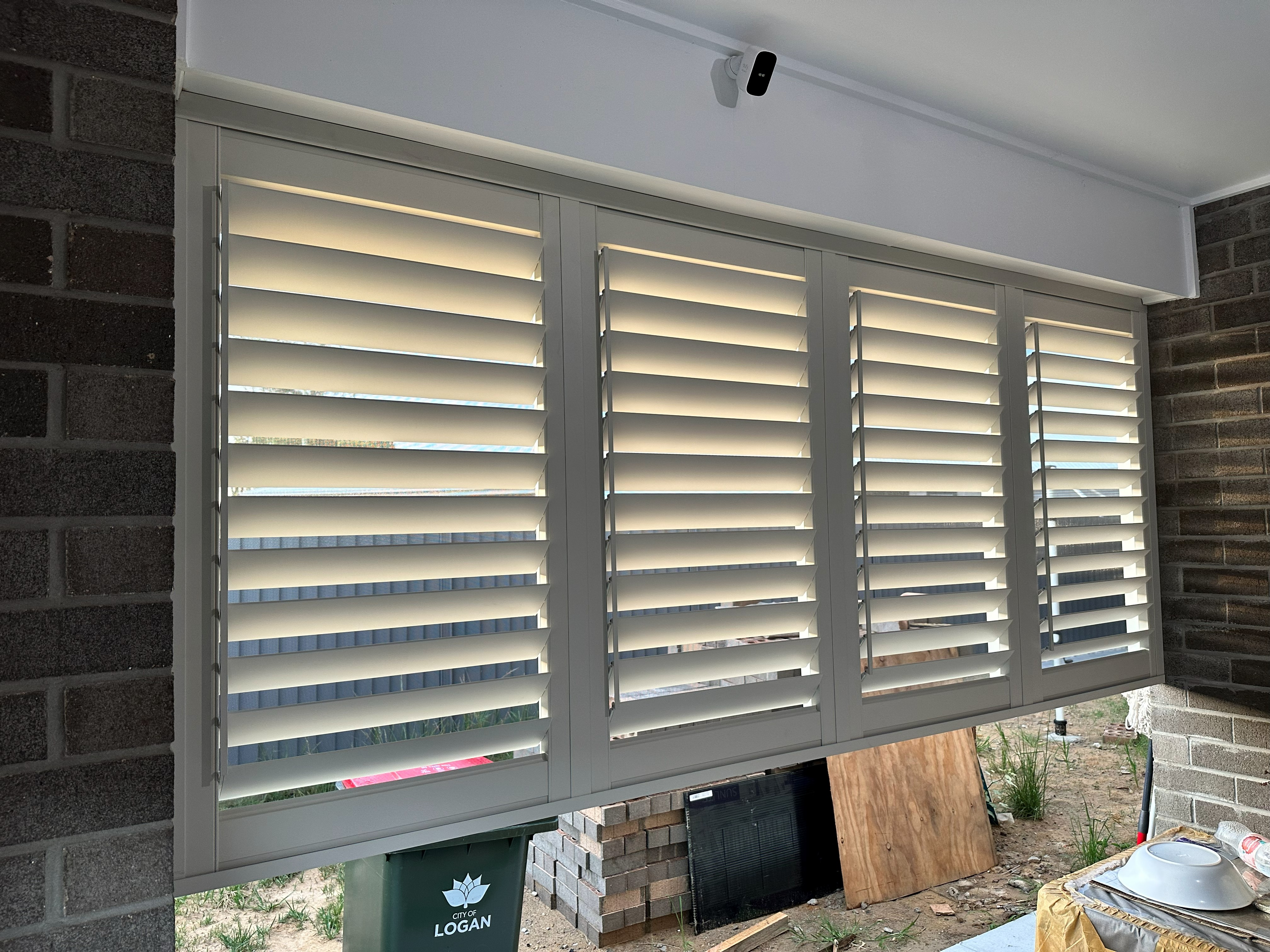 Grey aluminium shutters fitted to a brick home window with security camera above