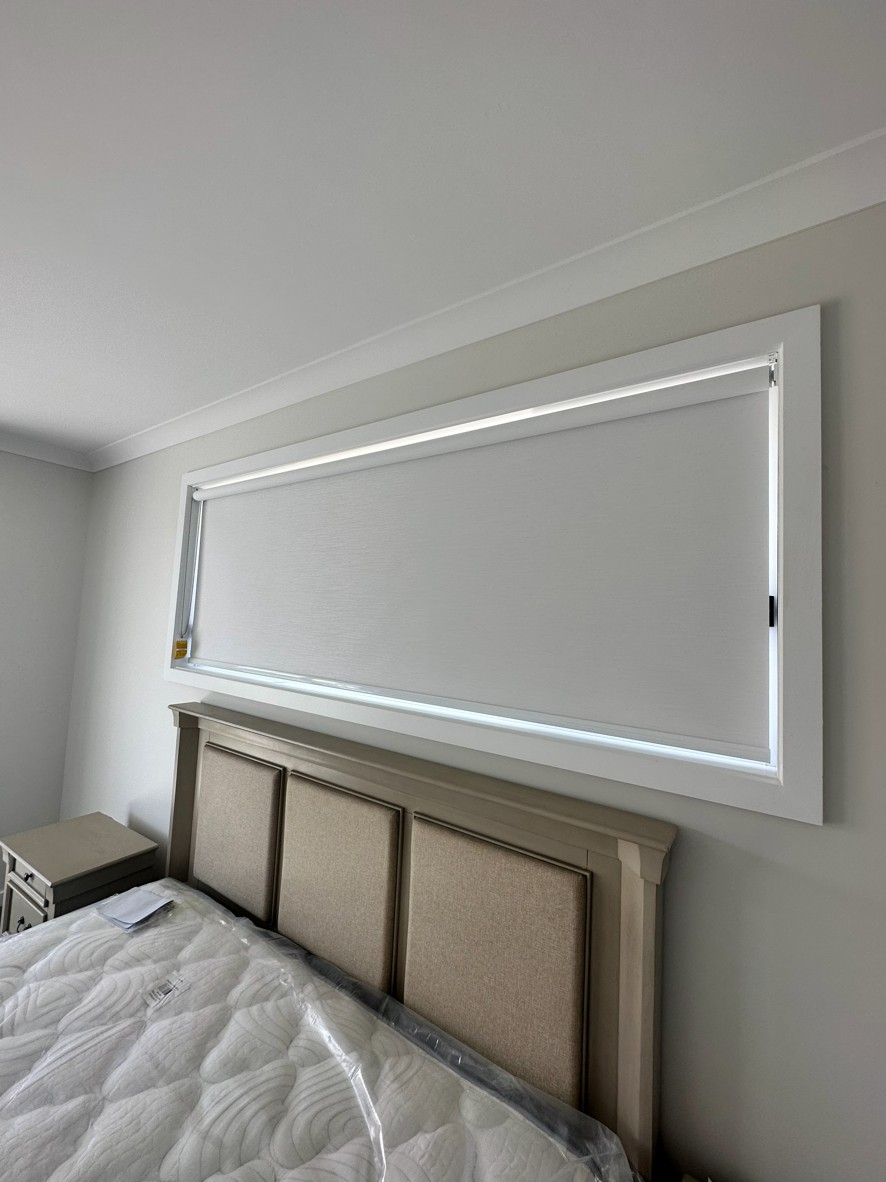 Light grey roller blind fully lowered over a bedroom window above a bed