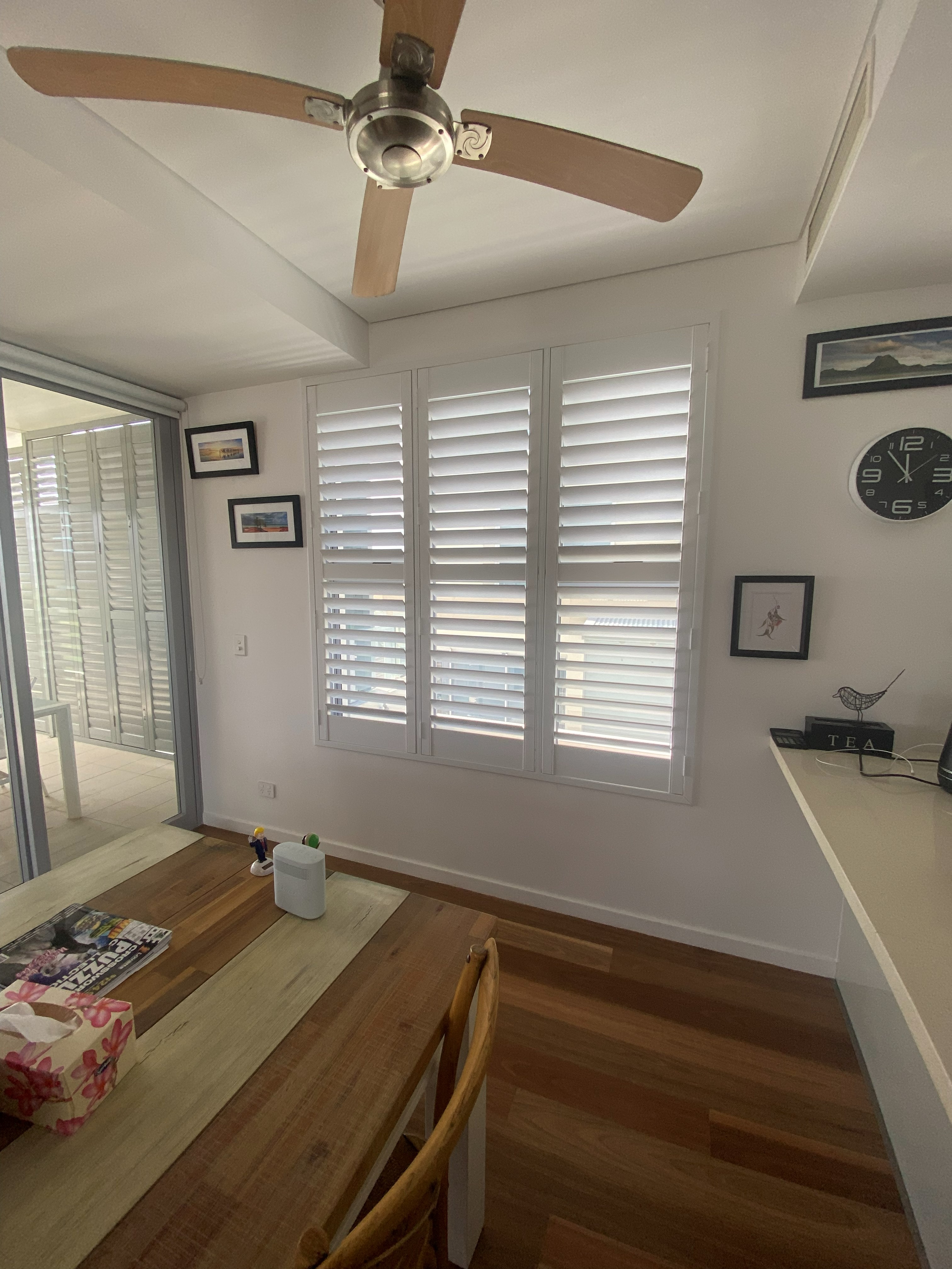 White plantation shutters in a dining area with ceiling fan and framed art
