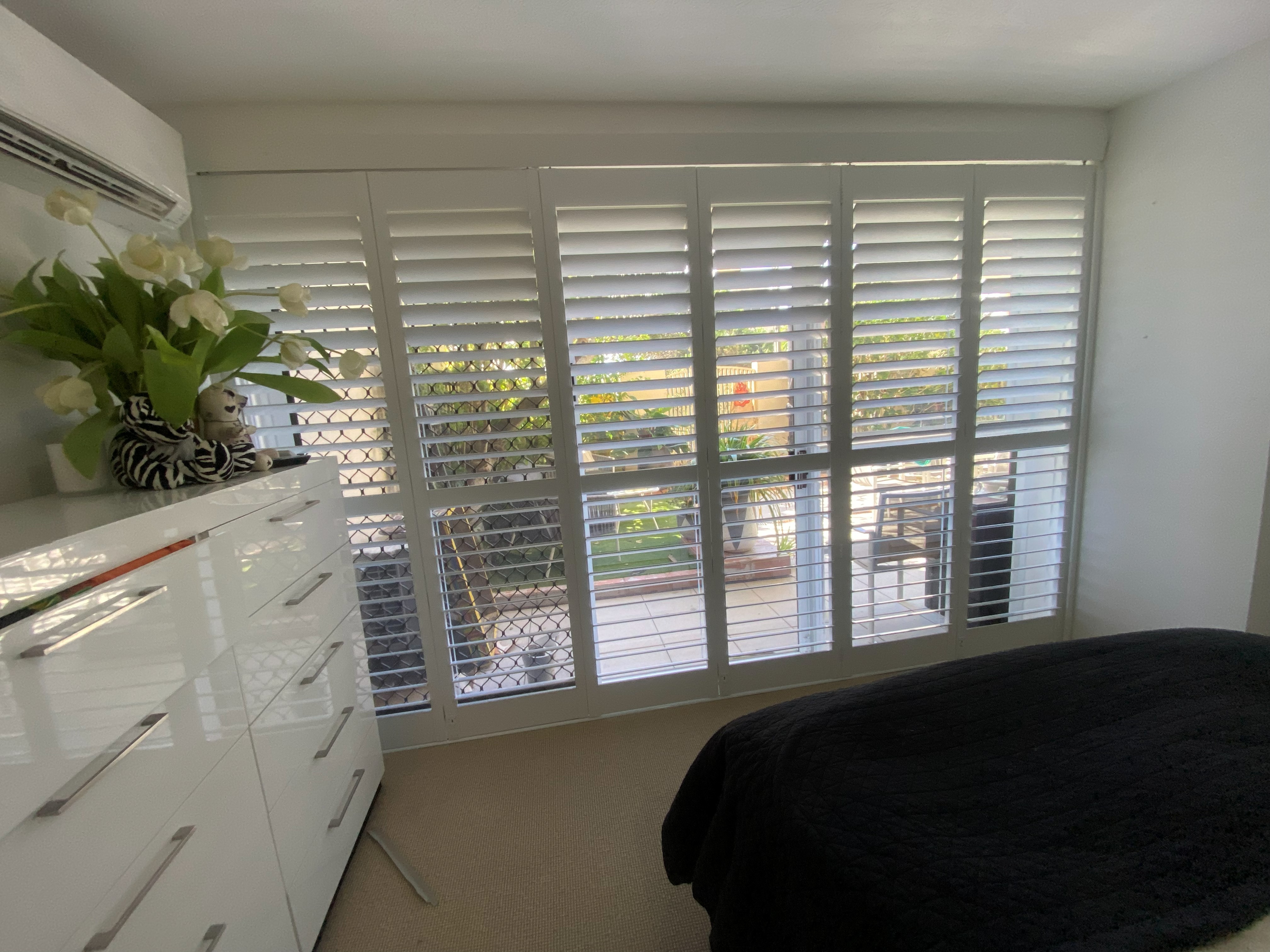 Plantation shutters in a bedroom with a dresser, flowers and pool view