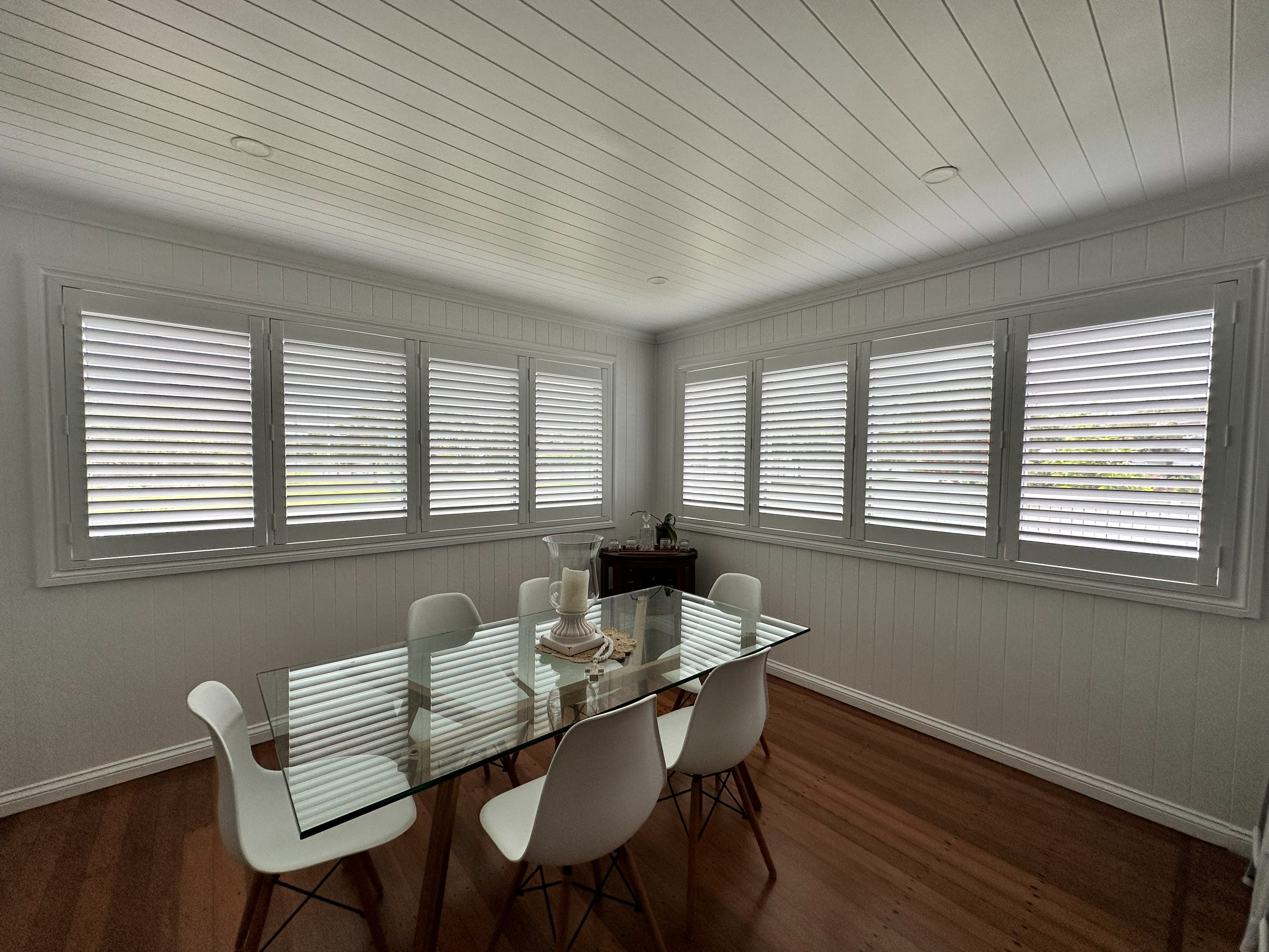 Corner plantation shutters in a dining room with a glass table and white chairs