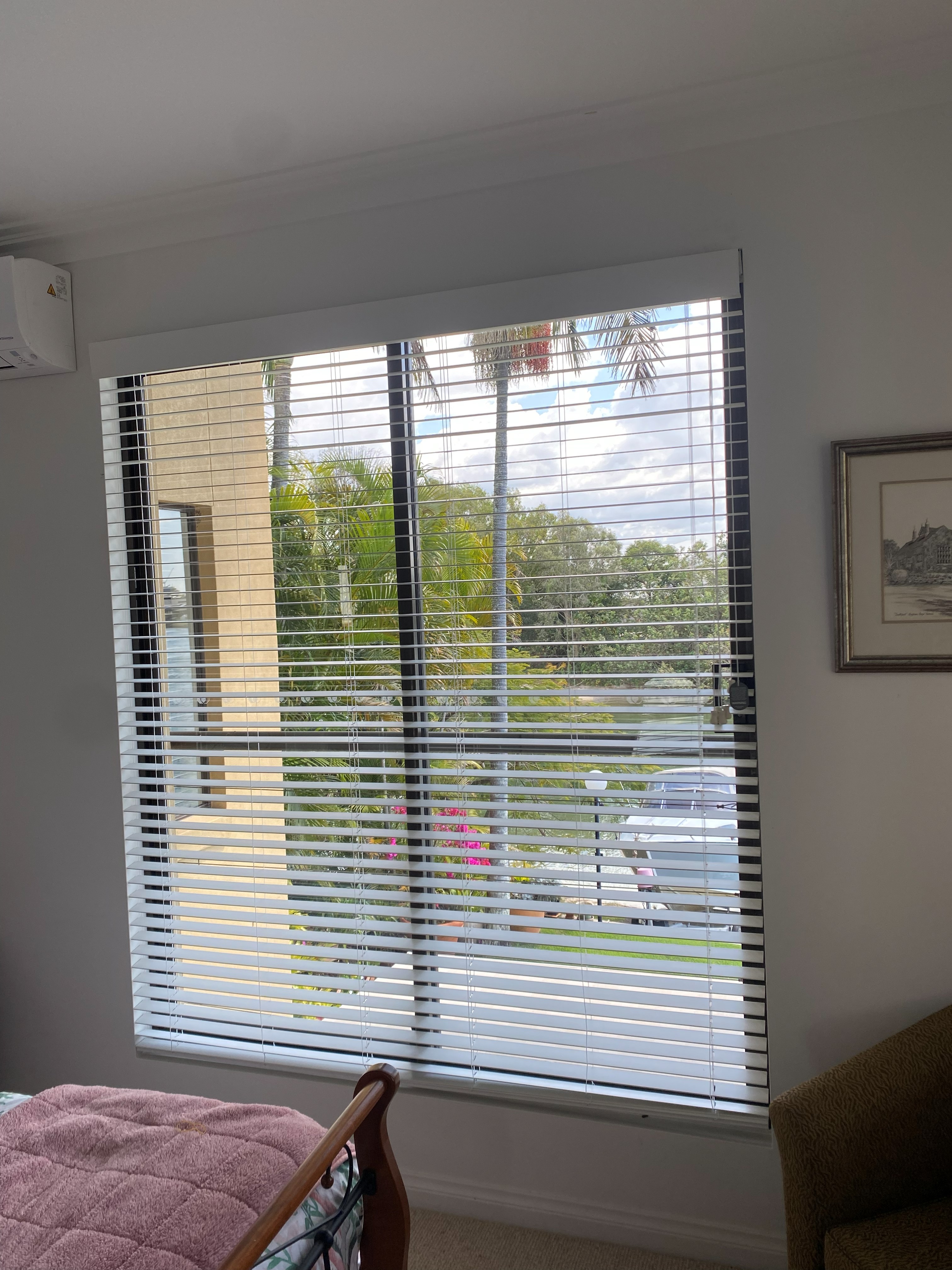 White venetian blinds fitted to a wide bedroom window with palm tree view